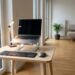Desk setup with a silver laptop on a stand, external keyboard and mouse on a light wood table by large glass doors.