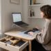 Woman sits at a wooden desk in a bright home office, using a laptop on a stand with a separate keyboard and mouse.