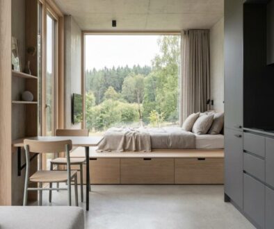 Open-plan studio with a wooden bed platform facing a forest view through large windows; kitchenette to the right.