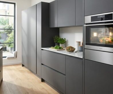 Modern gray kitchen with built-in stainless steel oven and tall cabinets; light wood floor and marble-topped island by a large window