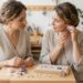 Two women in beige tops sit at a wooden table, crafting and comparing handmade jewelry with purple stones in a bright workshop.