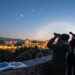 Group of four people on a stone viewpoint, gazing through binoculars at the Alhambra glowing at dusk, with a telescope, an open astronomy book, and a compass on the wall.