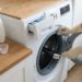 Person wearing gloves cleaning a front-loading washing machine with a brush at the door, detergent jar and spray bottle nearby on a wooden counter.