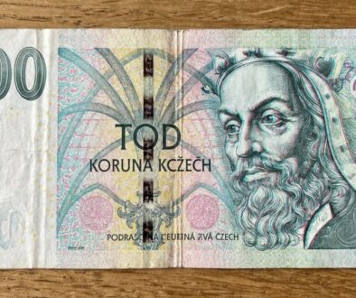 Front of a 100 Czech koruna banknote laid on a wooden surface, featuring a bearded man portrait and colorful decorative patterns.