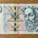 Front of a 100 Czech koruna banknote laid on a wooden surface, featuring a bearded man portrait and colorful decorative patterns.