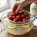 Hands hold a metal strainer full of fresh strawberries over a glass bowl of water with lemon slices and herbs inside a wooden counter kitchen scene.