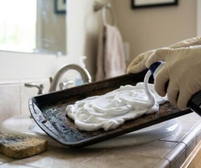 Person wearing gloves spraying foamy cleaner onto a baking sheet over a kitchen sink. The tray is being prepped for washing with a sponge and brush nearby.