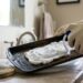 Person wearing gloves spraying foamy cleaner onto a baking sheet over a kitchen sink. The tray is being prepped for washing with a sponge and brush nearby.