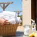 Person carrying a wicker laundry basket filled with white linens on a wooden table outdoors, with a clothesline in the background and a doorway to a house. A jar labeled Baking Soda, a bottle labeled Vinegar, and lemons on the table suggest natural cleaning supplies.