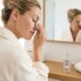 Woman in a white bathrobe wipes her eye with a cotton pad in front of a bathroom mirror.