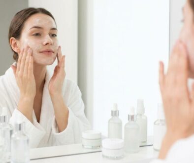 Woman in a white bathrobe applying facial cream while looking in a mirror, surrounded by skincare bottles on the counter