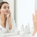 Woman in a white bathrobe applying facial cream while looking in a mirror, surrounded by skincare bottles on the counter