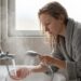 Woman in a gray bathrobe rinsing her hands under running water from a handheld shower at a tiled bathroom sink, near a window.