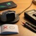 Smartwatch on a wireless charger with a nearby charging phone, an open notebook showing a battery icon and clock, and a digital multimeter with probes on a desk.