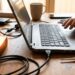 Hand on a silver laptop keyboard at a wooden desk, with tangled cables, an external drive, a digital thermometer, and a mug in the background.
