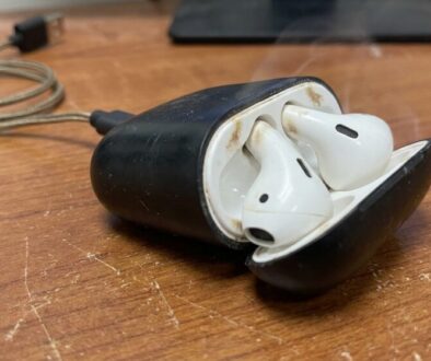White wireless earbuds (AirPods) in an open black charging case on a wooden desk.