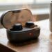 Wireless earbuds in an open charging case with a red status light on a wooden table beside a steaming mug.