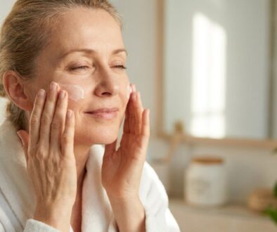 Older woman in a white bathrobe applying moisturizer to her cheeks in a bright bathroom with a mirror behind her.