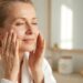 Older woman in a white bathrobe applying moisturizer to her cheeks in a bright bathroom with a mirror behind her.
