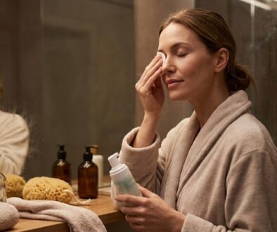 Woman in a beige bathrobe applying skincare near a bathroom mirror, holding a pump bottle.
