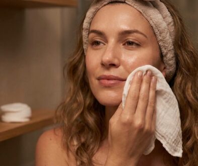 Woman with a towel headband wipes her cheek with a cloth during a skincare routine, with micellar water on the shelf in the background.