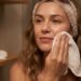 Woman with a towel headband wipes her cheek with a cloth during a skincare routine, with micellar water on the shelf in the background.