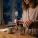 Person in a cozy kitchen pours a liquid from a bottle into a glass, with a mug, notebook, honey jar, and wooden counter nearby.