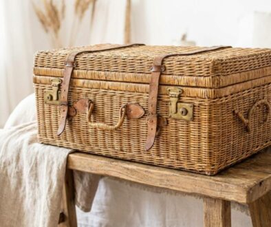 Woven wicker vintage suitcase with leather straps on a rustic wooden bench in a bright, neutral bedroom.