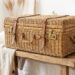 Woven wicker vintage suitcase with leather straps on a rustic wooden bench in a bright, neutral bedroom.