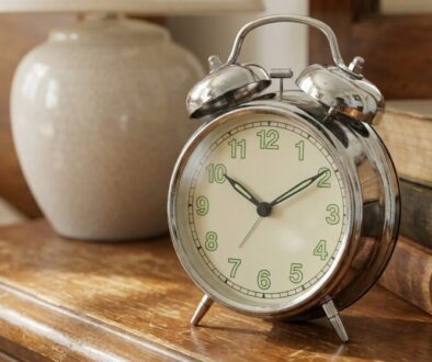 Vintage metal alarm clock on a wooden table beside a ceramic vase and a stack of old books.