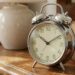 Vintage metal alarm clock on a wooden table beside a ceramic vase and a stack of old books.
