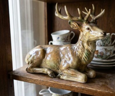 Ceramic deer figurine on a wooden shelf with porcelain teacups and plates in the background.