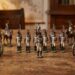 Miniature military figures arranged in rows with two small cannons on a wooden table, as if a tabletop display