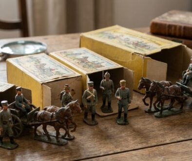 Diorama of vintage toy soldiers and horse-drawn carriages on a wooden table beside yellow boxes and a magnifying glass left edge