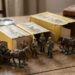 Diorama of vintage toy soldiers and horse-drawn carriages on a wooden table beside yellow boxes and a magnifying glass left edge