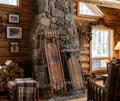 Cozy log-cabin living room with a stone fireplace, stacked firewood, and vintage wooden sleds beside a plaid stool and plaid throw on a chair.
