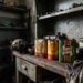 Dusty, cluttered mechanic's workshop with a weathered wooden workbench, rusty oil cans labeled Shell and Castrol, and dirty rags scattered about.