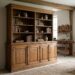 Rustic living room with a large wooden cabinet filled with books and pottery, a workbench against the wall, and a leather armchair on woven rugs.