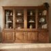 Large wooden china cabinet with glass doors in a sunlit living room, flanked by potted plants.