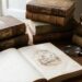 Stack of old leather-bound books on a wooden desk beside an open sketchbook with a vintage illustration, a magnifying glass, and a brass lamp by a window.