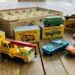 Assorted vintage Matchbox toy cars and a yellow crane truck on a wooden table with boxed sets in the background.