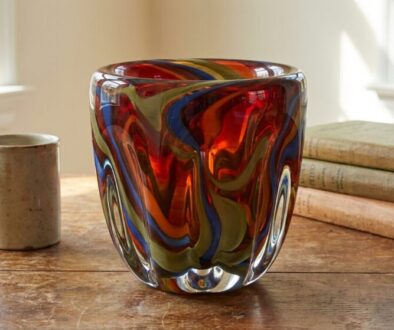 Colorful swirled glass vase on a wooden table beside a ceramic mug and a stack of old books by a window.