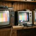 Three vintage CRT televisions on a wooden shelf displaying color test patterns from left to right: Grundig, Sony, and Tesla.