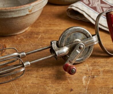 Vintage manual hand mixer with metal beaters on a wooden kitchen table, beside a ceramic bowl and patterned towel.