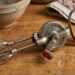 Vintage manual hand mixer with metal beaters on a wooden kitchen table, beside a ceramic bowl and patterned towel.
