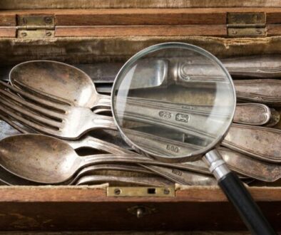 Open wooden box filled with tarnished silverware (spoons and forks); a magnifying glass rests on top, inspecting the utensils for patina or marks.