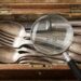 Open wooden box filled with tarnished silverware (spoons and forks); a magnifying glass rests on top, inspecting the utensils for patina or marks.