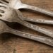 Group of tarnished metal forks with decorative handles laid on a rustic wooden surface, showing wear and patina.