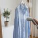 Person using a handheld garment steamer on a light blue shirt hanging on a rack, in a bright room with a plant nearby.