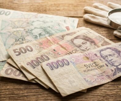 Several Czech koruna banknotes (100, 500, 1000 CZK) spread on a wooden table next to a magnifying glass and white gloves, suggesting money examination or counting.
